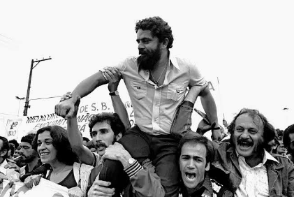 Lula, de barba e camisa clara, carregado nos ombros por metalúrgicos sorridentes, com faixas do sindicato ao fundo